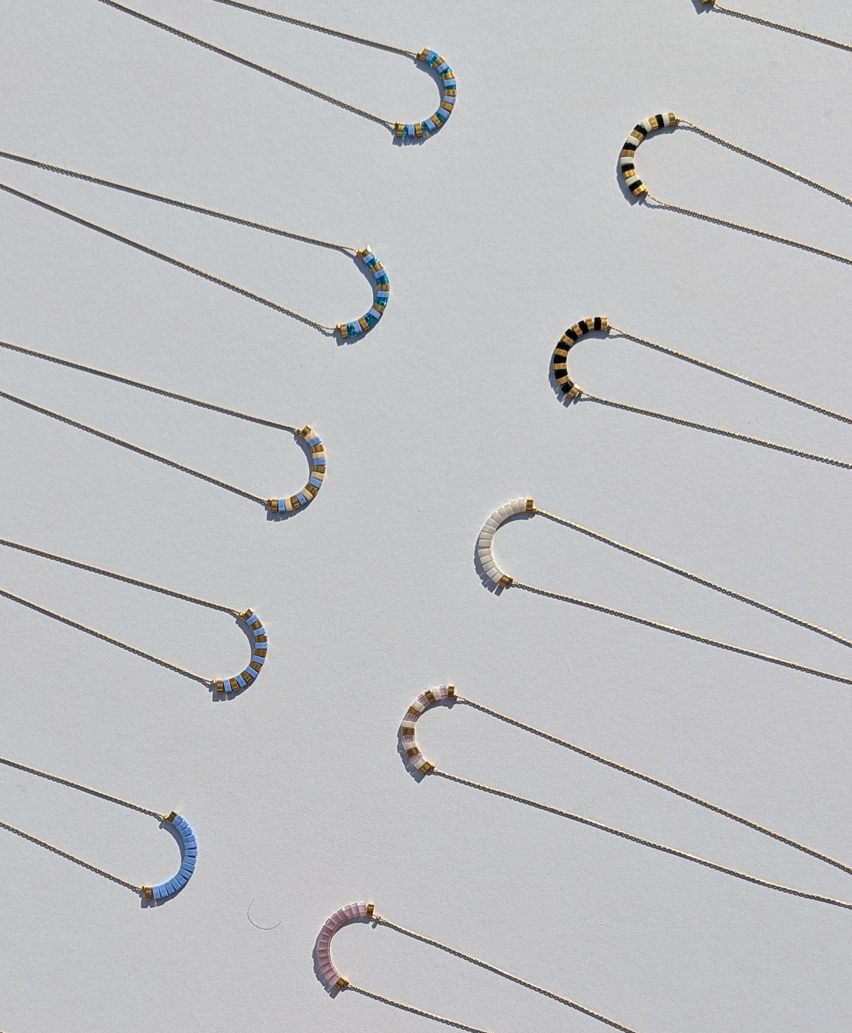 A white display sheet laid outdoors on a light surface, showing multiple gold-tone style necklaces with small bead accents in various colours including blue, green, pink, black, white and red.